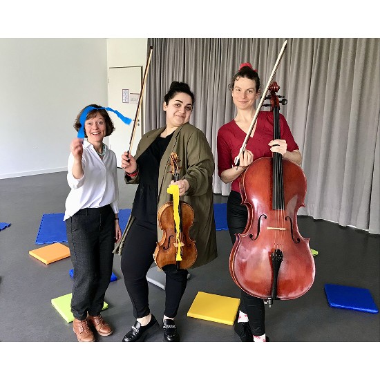 Drei Lehrerinnen stehend. Eine wedelt mit einem blauene Band eine hältihre Geige und eine ihr Cello