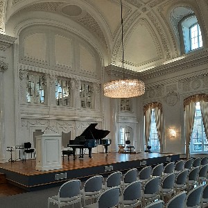 Foto des Weißen Saals: Eine Bühne mit Podium, schwarzem Flügel und kristallenem Lüster. Die Wände sind hoch, weiß und verziert.