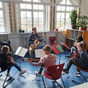 Ein Gitarrenlehrer und 4 Schüler und Schülerinnen spielen sitzend Gitarre