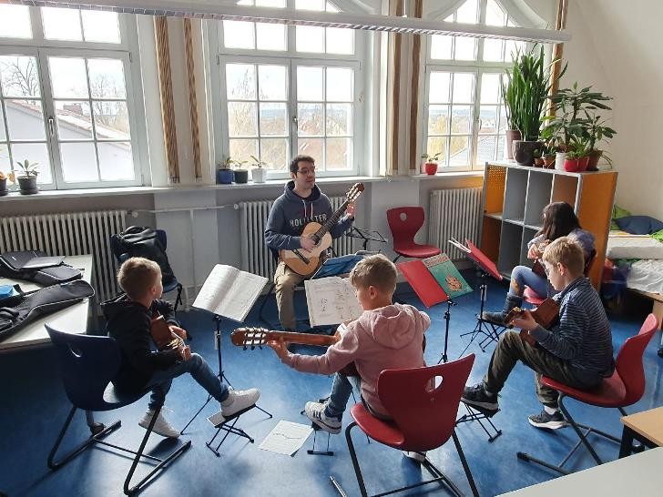 Lehrer spielt mit einer Schülerin und drei Schülern Gitarre. Alle sitzen.