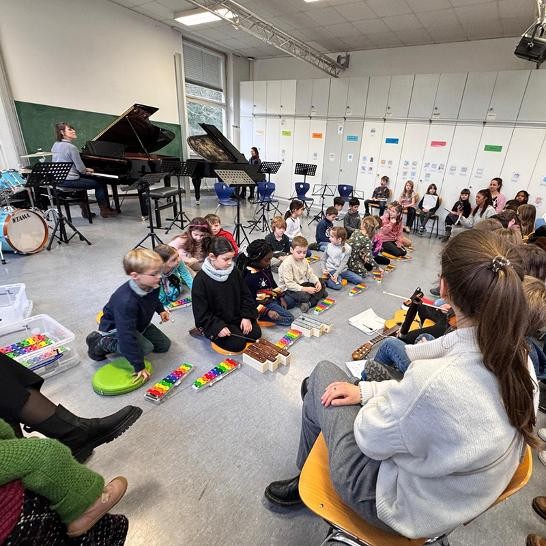 2 Lehrerinnen spielen am Flügel, viele Kinder sitzen davor am Boden und spielen Xylophon