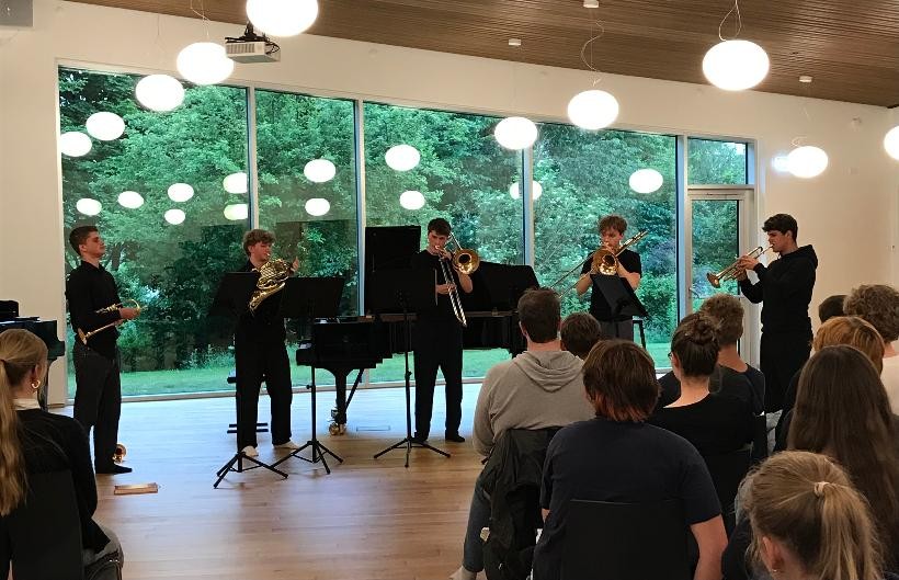 Ein Blechbläserquintett, 2 Trompeter, 2 Posaunisten, 1 Hornist, stehen in schwarzer Konzertkleidung in einem Konzertsaal und spielen gemeinsam. Das Foto wurde aus dem Publikum heraus aufgenommen.