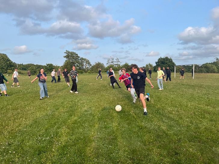 Ein Junge in schwarzem Fußballtrikot stürmt auf den Fußball zu und setzt zum Schuss an, andere Jugendliches stehen um ihn herum und versuchen den Ball zu erreichen