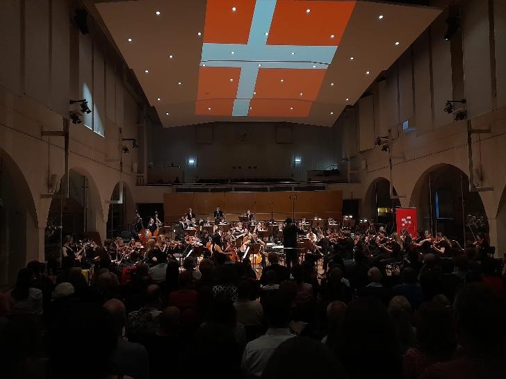 Jugendorchester sitzt auf der Bühne, der Konzertsaal ist dunkel. Das Foto wurde aus der letzten Publikumsreihe aufgenommen und die dänische Flagge ist an die Saaldecke projeziert