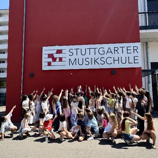 viele Kinder stehen mit dem Rücken zum Zuschauer und zeigen mit dem Finger auf eine rote Wand mit dem Musikschullogo