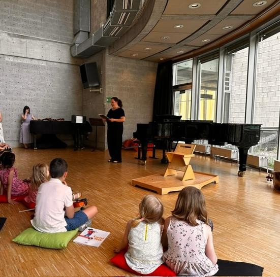 Kinder sitzen auf dem Boden in einem Saal, Lehrerin steht