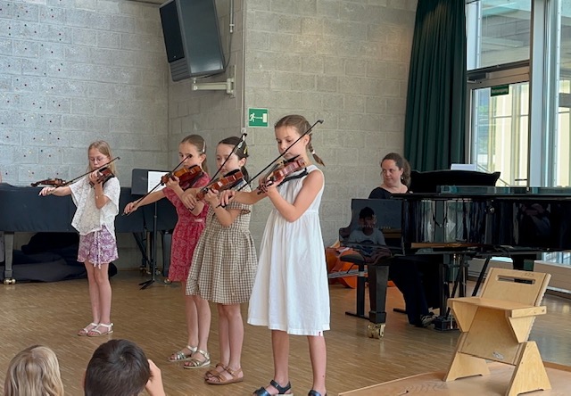 Lehrerin spielt Flügel. 4 Mädchen stehen davor und spielen Geige